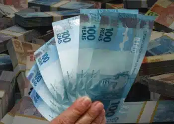 Mão segurando notas de R$ 100 em frente a pilhas de dinheiro brasileiro
