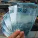 Mão segurando notas de R$ 100 em frente a pilhas de dinheiro brasileiro