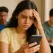 Estudante com expressão preocupada observa o celular, mão na cabeça, colegas ao fundo