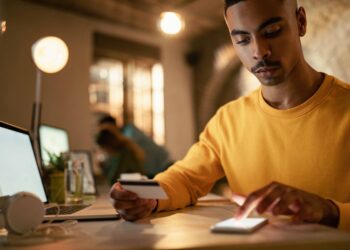 Homem com cartão de crédito usando smartphone ao lado de notebook em ambiente interno