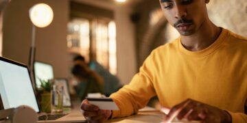 Homem com cartão de crédito usando smartphone ao lado de notebook em ambiente interno