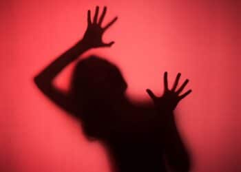 Silhueta escura e borrada de uma pessoa com as mãos levantadas e abertas em desespero e medo, pressionando contra uma tela ou pano vermelho brilhante e texturizado.