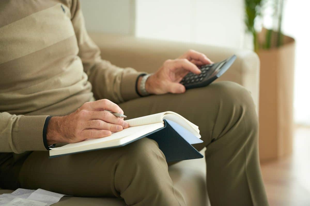 Homem faz cálculos financeiros com caderno e calculadora no sofá.