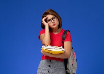 Jovem de camiseta vermelha, óculos e mochila cinza segura livros e toca a testa com expressão cansada.