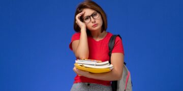 Jovem de camiseta vermelha, óculos e mochila cinza segura livros e toca a testa com expressão cansada.