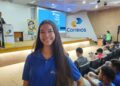 Jovem mulher sorridente, com cabelos pretos e longos, em pé no corredor de um auditório lotado. Ela veste uma camisa polo azul royal com o logotipo dos Correios no peito esquerdo. O auditório está cheio de pessoas sentadas nas poltronas brancas e azuis. No palco, ao fundo, um homem está no púlpito fazendo uma apresentação e um telão exibe o logotipo dos Correios e informações sobre o programa Jovem Aprendiz. Há também um banner no palco com a identidade visual do programa. A iluminação é de ambiente de conferência, simbolizando o Jovem Aprendiz dos Correios.