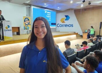 Jovem mulher sorridente, com cabelos pretos e longos, em pé no corredor de um auditório lotado. Ela veste uma camisa polo azul royal com o logotipo dos Correios no peito esquerdo. O auditório está cheio de pessoas sentadas nas poltronas brancas e azuis. No palco, ao fundo, um homem está no púlpito fazendo uma apresentação e um telão exibe o logotipo dos Correios e informações sobre o programa Jovem Aprendiz. Há também um banner no palco com a identidade visual do programa. A iluminação é de ambiente de conferência, simbolizando o Jovem Aprendiz dos Correios.
