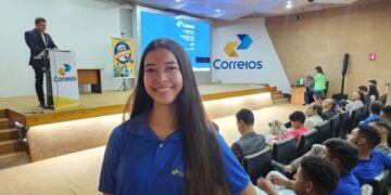 Jovem mulher sorridente, com cabelos pretos e longos, em pé no corredor de um auditório lotado. Ela veste uma camisa polo azul royal com o logotipo dos Correios no peito esquerdo. O auditório está cheio de pessoas sentadas nas poltronas brancas e azuis. No palco, ao fundo, um homem está no púlpito fazendo uma apresentação e um telão exibe o logotipo dos Correios e informações sobre o programa Jovem Aprendiz. Há também um banner no palco com a identidade visual do programa. A iluminação é de ambiente de conferência, simbolizando o Jovem Aprendiz dos Correios.