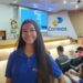Jovem mulher sorridente, com cabelos pretos e longos, em pé no corredor de um auditório lotado. Ela veste uma camisa polo azul royal com o logotipo dos Correios no peito esquerdo. O auditório está cheio de pessoas sentadas nas poltronas brancas e azuis. No palco, ao fundo, um homem está no púlpito fazendo uma apresentação e um telão exibe o logotipo dos Correios e informações sobre o programa Jovem Aprendiz. Há também um banner no palco com a identidade visual do programa. A iluminação é de ambiente de conferência, simbolizando o Jovem Aprendiz dos Correios.