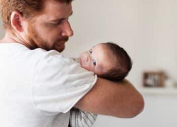 Homem adulto com barba ruiva segura bebê de cabelos escuros no colo, ambiente interno claro.