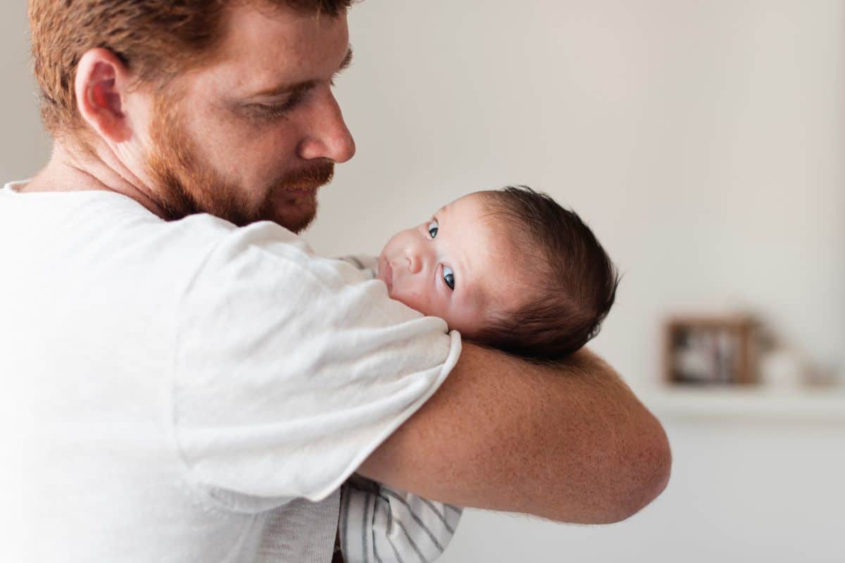 Homem adulto com barba ruiva segura bebê de cabelos escuros no colo, ambiente interno claro.