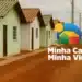 Conjunto de casas populares com paredes claras e telhados cerâmicos ao lado de rua de terra, logo Minha Casa Minha Vida.