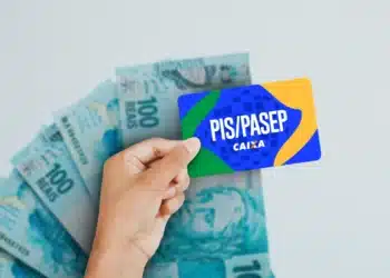 Mão segurando cartão PIS/PASEP azul, verde e amarelo da Caixa com notas de R$100 ao fundo.