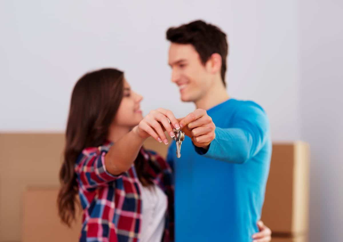 Casal jovem sorrindo juntos enquanto segura chave de casa em destaque, caixas de mudança ao fundo.