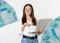 Jovem estudante com laptop cercada por notas de 100 reais.