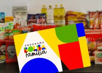 Cartão do Programa Bolsa Família colorido posicionado sobre mesa, com garrafas de óleo e alimentos embalados ao fundo.