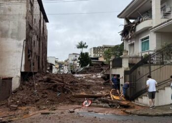 Rua em Minas Gerais obstruída por grande volume de lama, entulho e troncos de árvores após enchentes, com casas danificadas e moradores observando os estragos.