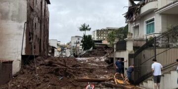 Rua em Minas Gerais obstruída por grande volume de lama, entulho e troncos de árvores após enchentes, com casas danificadas e moradores observando os estragos.