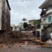 Rua em Minas Gerais obstruída por grande volume de lama, entulho e troncos de árvores após enchentes, com casas danificadas e moradores observando os estragos.