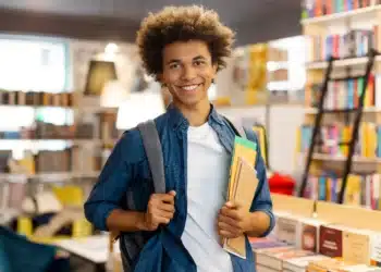 Jovem estudante negro com cabelo afro sorrindo, utilizando mochila e segurando cadernos coloridos em biblioteca.