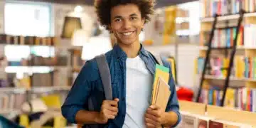 Jovem estudante negro com cabelo afro sorrindo, utilizando mochila e segurando cadernos coloridos em biblioteca.