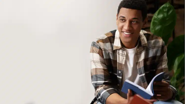 Diferença do programa Todo Jovem na Escola e o Pé-de-Meia Jovem negro sorridente segurando livro azul aberto, vestindo camisa xadrez marrom e bege, fundo claro com planta verde.
