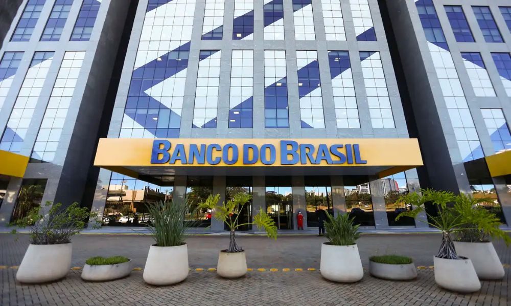 Fachada moderna de um grande edifício corporativo com paredes espelhadas refletindo o céu. Acima da entrada principal, um letreiro amarelo grande e proeminente com a inscrição "BANCO DO BRASIL" em letras azuis. No primeiro plano, há vasos de plantas ornamentais em uma calçada de paralelepípedos.
