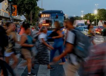 Pessoas atravessando faixa de pedestres com ônibus ao fundo em dia movimentado