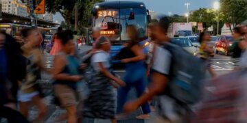 Pessoas atravessando faixa de pedestres com ônibus ao fundo em dia movimentado