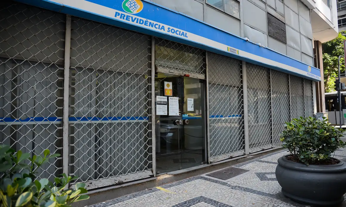 Agência da Previdência Social do INSS com grades metálicas Fachada de agência da Previdência Social com letreiro azul, grades metálicas fechadas e porta de vidro com avisos