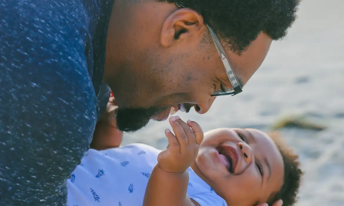 Lei sancionada em 2026 amplia licença-paternidade para até 20 dias e cria salário-paternidade pelo INSS Pai interagindo carinhosamente com bebê sorridente, representando a nova lei de licença-paternidade sancionada em 2026 que amplia o direito ao afastamento remunerado para pais trabalhadores