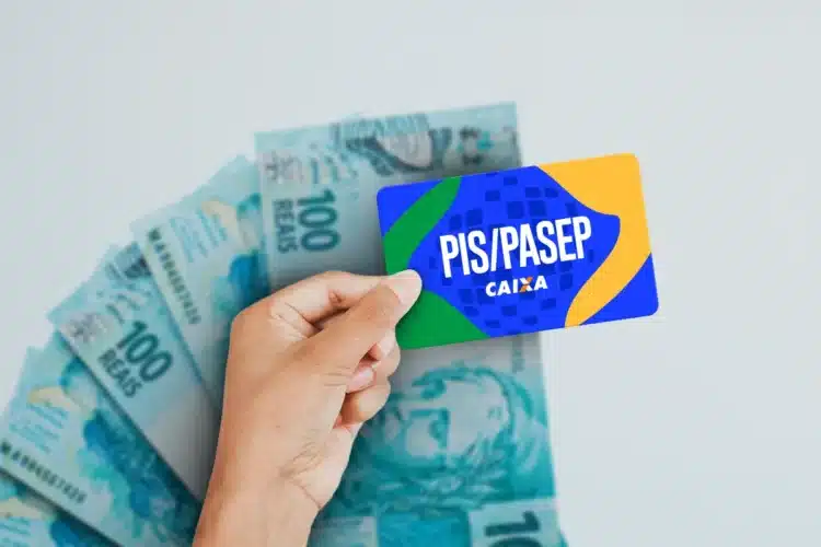 PIS/Pasep 2026 Mão segura cartão PIS/PASEP azul, verde e amarelo da Caixa sobre notas de R$ 100 ao fundo.