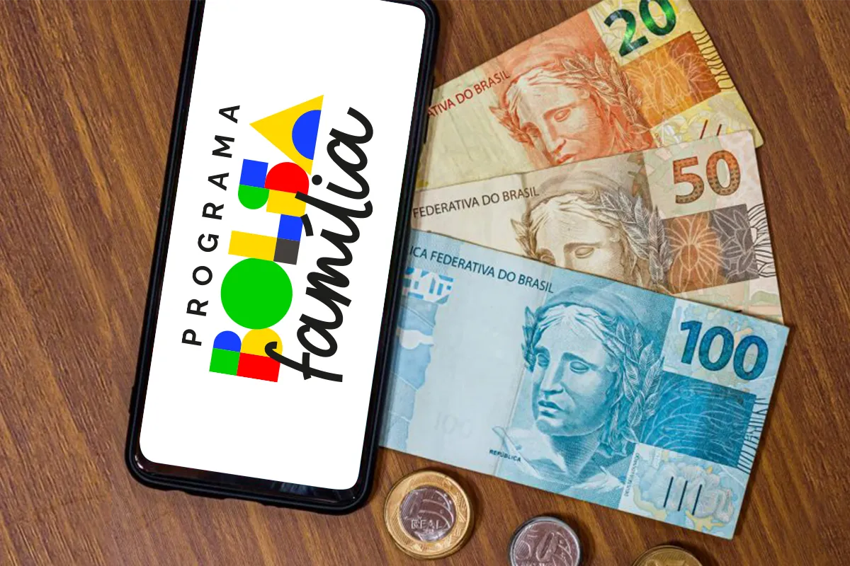 Bolsa família repasse financeiro app celular Foto em ângulo superior sobre mesa de madeira, mostrando um smartphone preto com a logomarca do Bolsa Família na tela. Ao lado, cédulas de Real (R$ 100, R$ 50 e R$ 20) e moedas, simbolizando o repasse mensal e o pagamento do benefício.