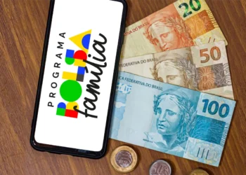 Smartphone exibindo logo do Programa Bolsa Família ao lado de notas de R$100, R$50 e R$20 e moedas sobre superfície de madeira.