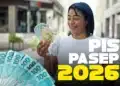 Mulher sorridente de camiseta branca segura notas de 100, 50 e 10 reais, com texto PIS Pasep 2026 ao lado.