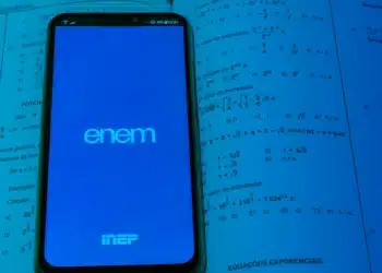 Smartphone exibindo logo do Enem e INEP em tela azul sobre páginas de livro de matemática com equações exponenciais.