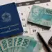 Carteira de trabalho azul, notas de 100 reais, calculadora, caneta preta e calendário de maio de 2026 sobre mesa clara