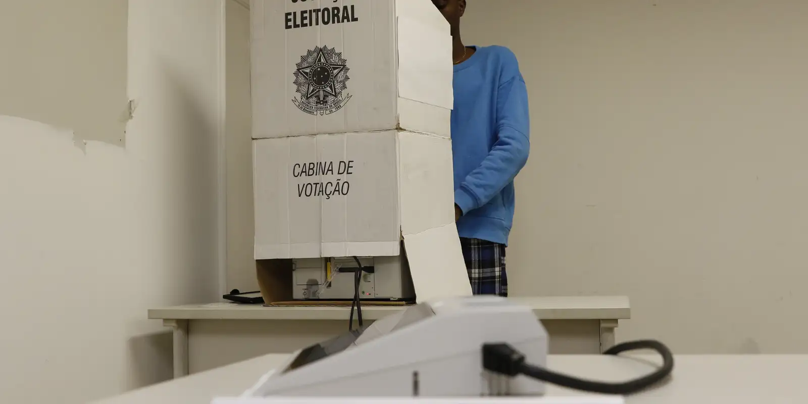 Eleitora vota em cabina de votação oficial da Justiça Eleitoral Brasileira Pessoa de blusa azul vota em cabina de votação com brasão e texto "ELEITORAL" e "CABINA DE VOTAÇÃO"