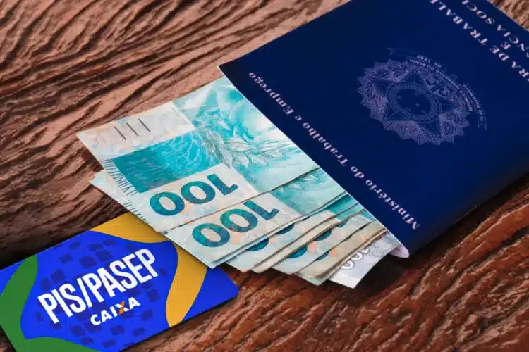 PIS/Pasep 2026 Carteira de trabalho azul sobre notas de R$ 100 reais ao lado de cartão PIS/PASEP da Caixa em superfície de madeira.