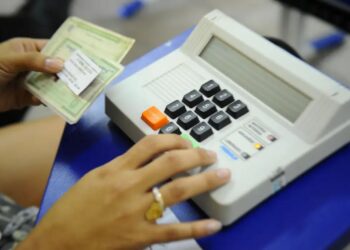 Close da mão de uma eleitora com anel dourado pressionando o botão verde "CONFIRMA" em uma urna eletrônica brasileira, enquanto a outra mão segura um documento impresso (título de eleitor).