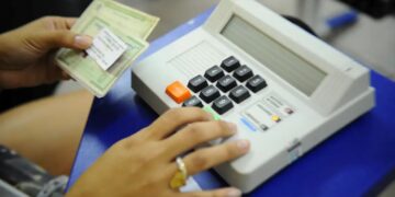 Close da mão de uma eleitora com anel dourado pressionando o botão verde "CONFIRMA" em uma urna eletrônica brasileira, enquanto a outra mão segura um documento impresso (título de eleitor).