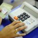 Close da mão de uma eleitora com anel dourado pressionando o botão verde "CONFIRMA" em uma urna eletrônica brasileira, enquanto a outra mão segura um documento impresso (título de eleitor).