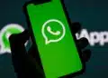 Mão segurando smartphone com logotipo do WhatsApp na tela verde, fundo com logo do aplicativo desfocado.