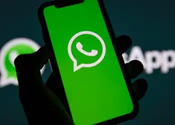 Mão segurando smartphone com logotipo do WhatsApp na tela verde, fundo com logo do aplicativo desfocado.