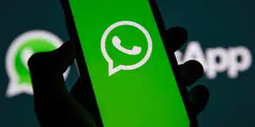 Mão segurando smartphone com logotipo do WhatsApp na tela verde, fundo com logo do aplicativo desfocado.