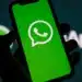 Mão segurando smartphone com logotipo do WhatsApp na tela verde, fundo com logo do aplicativo desfocado.