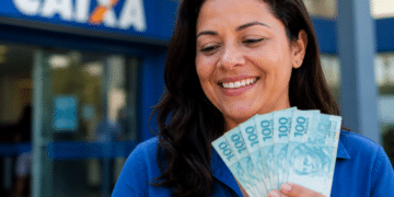 Mulher de blusa azul sorri segurando um leque de notas de R$ 100 em frente a uma agência da Caixa