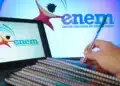 Notebook exibindo logo do Enem com mão segurando caneta sobre cadernos empilhados em ambiente de estudo