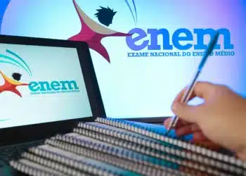 Notebook exibindo logo do Enem com mão segurando caneta sobre cadernos empilhados em ambiente de estudo