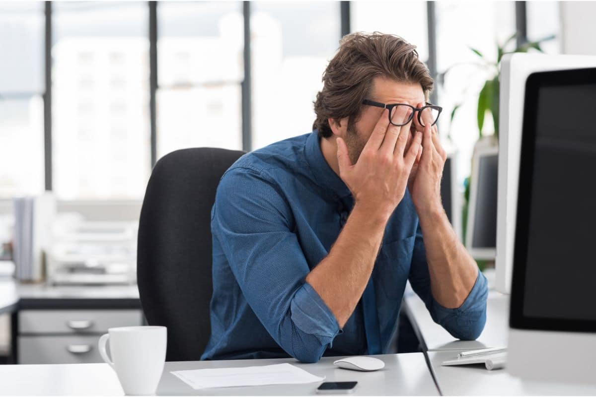 Homem de camisa azul e óculos esfrega os olhos sentado em frente ao computador em escritório iluminado